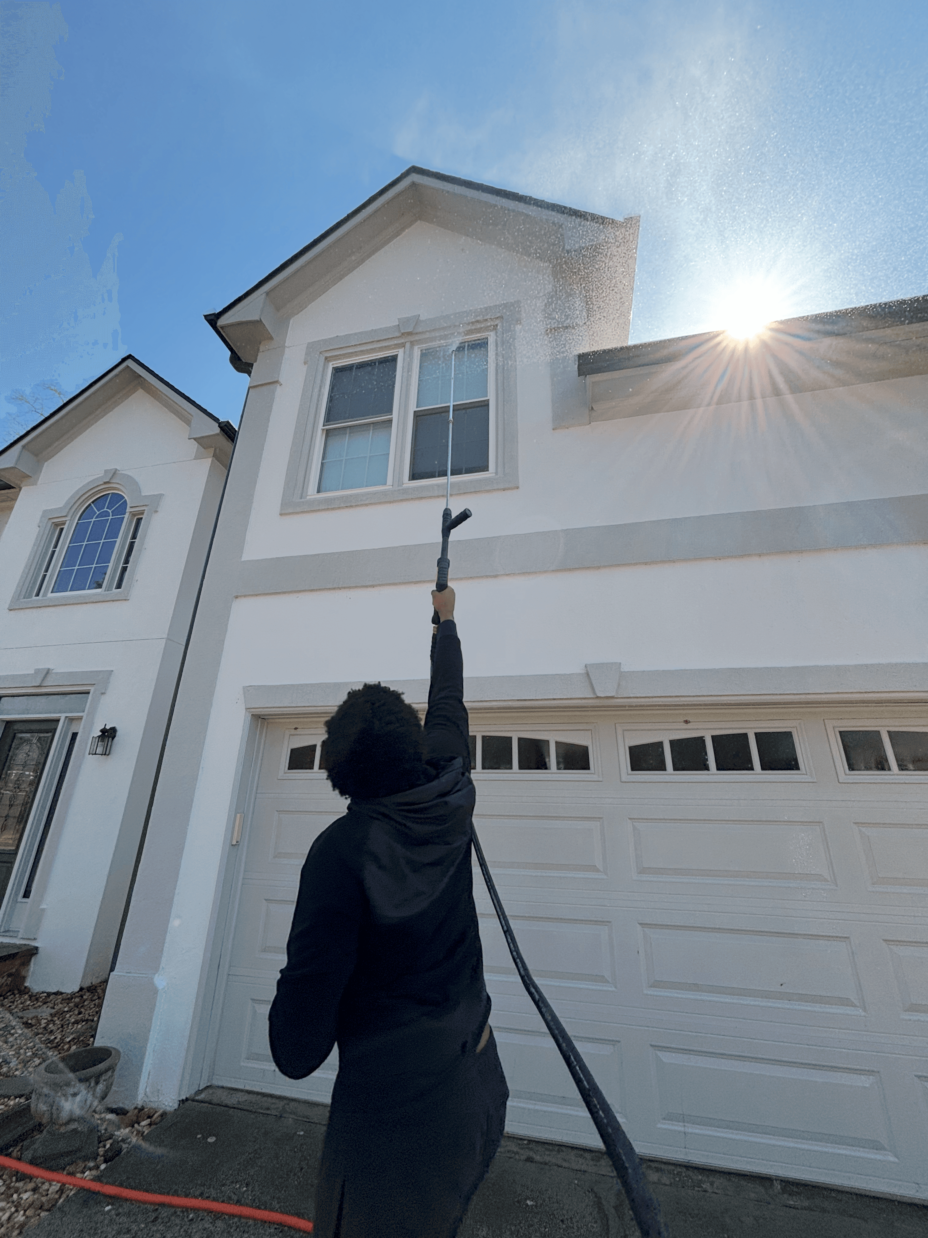 Person pressure washing a second-story window of a white house on a sunny day.